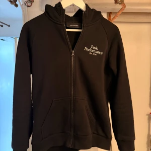 Svart hoodie från Peak Performance - Svart hoodie från Peak Performance med dragkedja och huva. Klassisk logga broderad på bröstet och stora fickor framtill. Skön passform och perfekt för lager-på-lager. Tillverkad i mjuk bomullsblandning.
