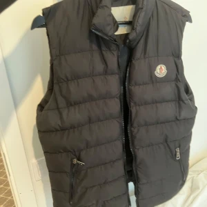 Svart dunväst från Moncler - Snygg svart dunväst från Moncler med klassisk logga på bröstet. Västen har quiltad design, hög krage och två fickor med dragkedja framtill. Perfekt att slänga över en hoodie eller tröja för extra värme. Stängs med dragkedja hela vägen upp.