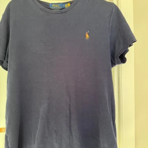 Blå t-shirt från Polo Ralph Lauren - Mörkblå t-shirt från Polo Ralph Lauren med klassisk rund hals och broderad logga på bröstet. Skön passform och tillverkad i mjuk bomull, perfekt till jeans eller shorts. Enkel och stilren design som funkar till det mesta.