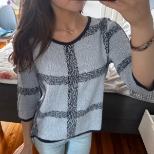 Grå rutig stickad tröja med trekvartsärm - Snygg grå stickad tröja med svart rutigt mönster och mörka kanter vid halsringning och ärmslut. Tröjan har trekvartsärmar och en avslappnad passform, perfekt till jeans för en chill look.