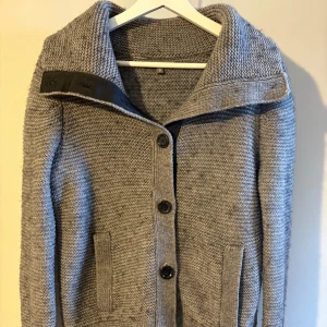 Grå stickad kofta med stora knappar - Mysig grå stickad cardigan med bred krage och stora svarta knappar framtill. Koftan har två fickor och en snygg detalj i svart vid kragen. Perfekt en stilren look!  