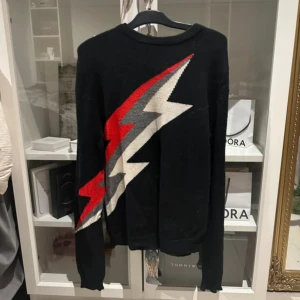 Svart tröja med blixt från Zadig & Voltaire - Cool svart stickad tröja från Zadig & Voltaire med en stor blixt i rött, grått och vitt över framsidan. Tröjan har rund halsringning och långa ärmar. Perfekt för dig som vill sticka ut med en edgy vibe.