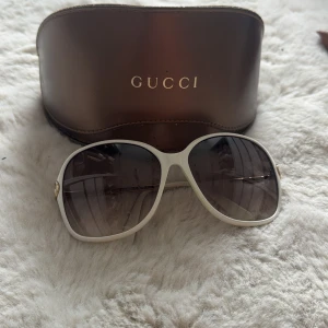 Vita solglasögon från Gucci - Snygga solglasögon från Gucci med vit båge och stora, lätt rundade glas. Sidorna har diskret Gucci-logga i guld. Kommer med original brunt fodral. Perfekta för dig som vill sticka ut med en lyxig accessoar.