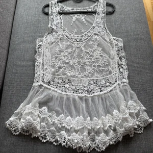 Vit spetsig topp med broderier. Nyskick. S - *Superfin vit topp i transparent mesh med broderade blommor och spetsdetaljer.  *Ärmlös modell med rund halsringning och volangkant nertill.  *Perfekt för dig som gillar romantisk och bohemisk stil. *Den har en lös passform och en vågig nederkant. *Etiketten är bortklippt. Storlek S *Mycket bra skick. Tvättad *Djurfritt och rökfritt hem