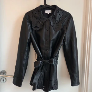 Svart skinnjacka & other stories  - Supersnygg feminin svart äkta skinnjacka från  Other Stories med unik laserskuren krage och nederkant. Jackan har knappar framtill, bälte i midjan och långa ärmar. Perfekt statement-plagg med coola detaljer som ger en edgy vibe. Oanvänd/ny. Nypris 3500 krage.