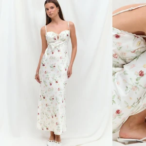 Vit blommig långklänning med spets - Romantisk långklänning i vit satin med blommigt mönster i rosa, grönt och rött. Klänningen har smala axelband, söt spetskant och knytning framtill vid bysten. Perfekt för sommarens alla tillfällen.