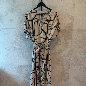 Beige och svart klänning stl S/M - Snygg beige från Lindex med svart abstrakt mönster. Klänningen har korta ärmar, knappar, knytband i midjan och v-ringning. Perfekt för dig som gillar en avslappnad men trendig stil. Luftig modell i härlig viskos. 