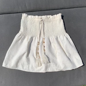 Vit topp med snörning med snäckor - Superfin vit topp i bomull och linne med snörning med snäckor och tofsar på. Toppen har en strukturerad väv och känns både luftig och trendig. Perfekt för varma dagar och ger en chill boho-vibe.