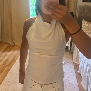 Vit halterneck topp - Säljer en stilren vit halterneck topp med draperad halsringning. Toppen är ärmlös och har en tight passform som framhäver figuren. Perfekt för dig som gillar minimalistisk och fräsch stil. Endast testad!