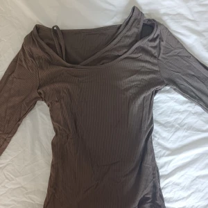 Brun ribbad långärmad topp Lewkin - Säljer en brun ribbad långärmad topp från Lewkin med en cool omlott-look och en extra axelbandsdetalj. Toppen har en figurnära passform och är gjord i mjuk polyester. Perfekt för dig som gillar trendiga och unika snitt!