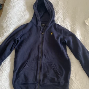 Mörkblå hoodie från Lyle & Scott - Snygg mörkblå hoodie från Lyle & Scott med dragkedja och huva. Klassisk gul logga på bröstet och två stora fickor framtill. Perfekt för chill dagar eller när du vill ha en enkel och stilren look.