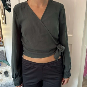 Mörkgrön omlottopp från BubbleRoom - Snygg mörkgrön omlottopp från BubbleRoom med långa ärmar och knyt i sidan. Toppen är croppad och har smockade muddar vid ärmsluten. Perfekt för dig som gillar stilrena och trendiga plagg med det lilla extra.