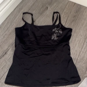 Svart linnetopp från First Position - Svart linnetopp från First Position, säljs endast på Lindex. Toppen har tunna axelband och en snygg broderad detalj med bladrankor på ena sidan. Materialet är mjukt och stretchigt, perfekt för en stilren look.