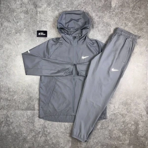 Grå träningsset från Nike - Snyggt grått träningsset från Nike med jacka och byxa. Jackan har huva, dragkedja och två fickor framtill. Byxorna har resår i midjan och diskret Nike-logga på benet. Perfekt för dig som gillar sportig stil.