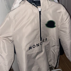 Rep Berge moncler jacka - Snygg beige vindjacka från Moncler med svart och grön logga på bröstet. Jackan har en stor ficka framtill med Moncler-tryck, halv dragkedja och svart innerfoder. Perfekt för dig som vill ha en stilren och sportig look.