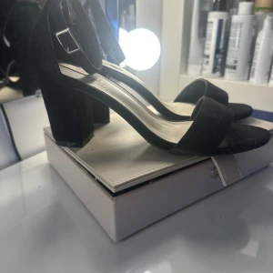 Svarta klackade sandaler från Nelly - Stilrena svarta sandaler från Nelly med bred klack och öppen tå. Skorna har en enkel rem över tårna och justerbart spänne vid ankeln. Ytan är i mockaliknande material som ger en lyxig känsla. Perfekta för att lyfta vilken outfit som helst. Endast använda en gång