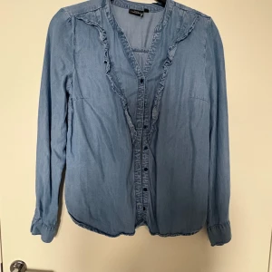 Blå volangskjorta från KappAhl - Supersöt ljusblå skjorta från KappAhl i mjuk bomull. Skjortan har långa ärmar, knappar framtill och fina volangdetaljer längs knappraden. Perfekt för dig som gillar en chill men ändå feminin stil.