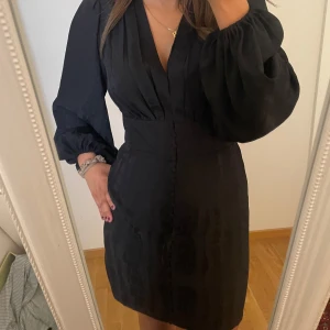 Svart kort klänning med puffärm - Supersnygg svart kort klänning med djup v-ringning markerad midja och puffiga långärmar. Efterliknar ADOORE klänningar🤩Klänningen har dekorativa knappar framtill och ett lätt glansigt tyg som ger en lyxig känsla. Perfekt för dig som vill sticka ut med stilrena detaljer. Den är från NA-KD, säljs tyvärr inte längre då den är köpt förra året men endast använd en gång!🤩