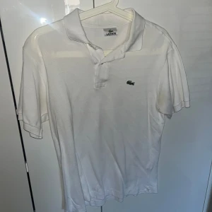 Vit pikétröja från Lacoste - Klassisk vit pikétröja från Lacoste med broderad grön krokodil på bröstet. Tröjan har korta ärmar, krage och knäppning framtill. Tillverkad i bomull för en skön och luftig känsla. Perfekt för en clean och sportig stil.