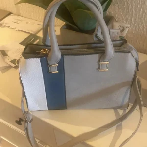 Grå och blå axelväska  - Snygg axelväska  i grått, blått och vitt med stilrena guldiga detaljer. Väskan har både handtag och justerbar axelrem samt flera fack inuti för smart organisering. Perfekt för dig som vill ha en praktisk och trendig väska med plats för allt du behöver.