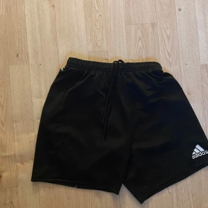 Svarta träningsshorts från Adidas - Svarta shorts från Adidas med klassisk logga på benet. De har elastisk midja med snörning och är tillverkade i ett lätt och sportigt material som passar perfekt för träning eller chill. Snygg och enkel design som funkar till det mesta.