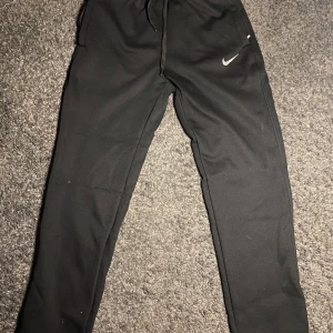 Svarta mjukisbyxor från Nike - Svarta mjukisbyxor från Nike med klassisk vit swoosh-logga på låret. Byxorna har dragsko i midjan och raka ben. Perfekta för chill eller träning, tillverkade i mjukt material som känns skönt mot huden. Passar som stolek S/XS