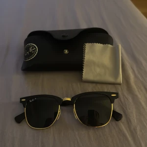 Ray-Ban Clubmaster solglasögon svart/guld - Snygga Ray-Ban Clubmaster solglasögon med svart ovandel och guldfärgade detaljer. Klassisk halvbåge och mörka polariserade glas. Kommer med originalfodral och putsduk. Perfekt accessoar för en cool och tidlös stil.