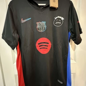 FC Barcelona svart Nike fotbollströja - Snygg svart FC Barcelona fotbollströja från Nike med röda och blå sidopaneler. Tröjan har klubbmärke, Nike-logga, Spotify-logga och ett vitt smiley-tryck på bröstet. Tillverkad i lätt och ventilerande material, perfekt för match eller träning.