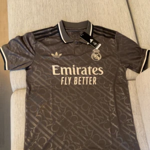 Real Madrid x Adidas grå fotbollströja - Snygg grå Real Madrid fotbollströja från Adidas med diskret mönster över hela tyget. Tröjan har korta ärmar, klassisk krage med vita detaljer och svarta Adidas-ränder på axlarna. Stort Emirates-tryck på bröstet och klubbmärke i vitt. Perfekt för dig som älskar fotboll och streetstyle.