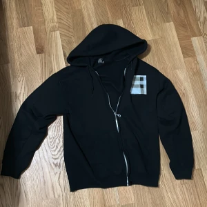 Svart hoodie med dragkedja och rutig detalj - Svart hoodie med hel dragkedja och huva. På bröstet finns en snygg rutig patch i grått, vitt och svart. Tröjan har långa ärmar, snörning i huvan och är tillverkad i mjukt bomullsmaterial. Perfekt för en chill och stilren look.