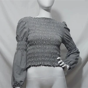 Grå smockad blus med puffärm - Trendig grå blus med smockad kropp och långa puffärmar. Modellen är croppad och har elastiska detaljer vid ärmslut och midja för en snygg passform. Perfekt för dig som gillar en stilren men ändå cool look.