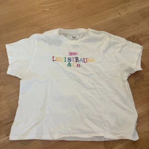 Vit t-shirt från Levi's med färgglatt tryck - Vit t-shirt från Levi's i bomull med färgglatt broderat 'Levi Strauss & Co.' på bröstet. Klassisk passform och korta ärmar, perfekt till jeans eller shorts. Enkel och stilren med en twist av färg.