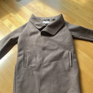 Brun kappa från Vila med bred krage - Säljer en stilren brun kappa från Vila med bred krage och raka fickor. Jackan har en minimalistisk design och är tillverkad i ett mjukt material som känns lyxigt. Perfekt för dig som gillar en clean och klassisk look. Saknar ett midjeband 