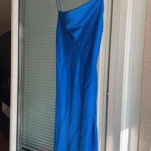 Blå långklänning från Astrid Frank - Supersnygg blå långklänning från Astrid Frank  med tunna axelband och slät, glansig yta. Klänningen har en enkel, elegant siluett och är perfekt för dig som gillar minimalistisk stil. Materialet känns svalt och följsamt mot kroppen. Liten skada på ena bandet, men går enkelt att laga!