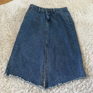 Blå jeanskjol från BluCollar, strl 28( M)  - Snygg blå lång jeanskjol från BluCollar med rå kant nertill och slits både fram och bak. Klassisk femficksmodell med bälteshällor och dragkedja. Gjord i kraftig denim med 97% bomull och 3% polyester. Perfekt för dig som gillar en chill och trendig look.