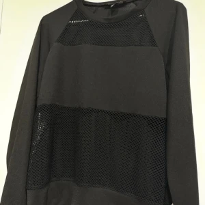 Svart långärmad tröja med meshdetaljer - Svart långärmad tröja med coola meshpaneler både upptill och nedtill på framsidan. Tröjan har en relaxed passform och rund halsringning, perfekt för en trendig och sportig look.