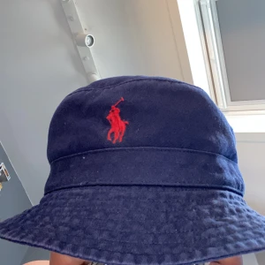 Mörkblå bucket hat från Polo Ralph Lauren - Snygg mörkblå bucket hat från Polo Ralph Lauren med klassisk röd broderad logga framtill. Hatten är tillverkad i 100% bomull . Perfekt för sommaren .