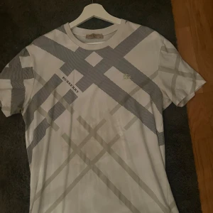 Vit rutig t-shirt från Burberry - Snygg vit t-shirt från Burberry med klassiskt rutmönster i grått och svart. T-shirten har korta ärmar och rund halsringning. Perfekt för dig som gillar stilrena och exklusiva plagg med ikonisk design.