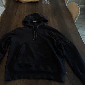 Svart hoodie från C.P. Company - Säljer en svart/mörkblå hoodie från C.P. Company med klassisk huva och känguruficka. Tröjan har en ikonisk linsdetalj på ärmen och ribbade muddar. Perfekt för en avslappnad och stilren look.