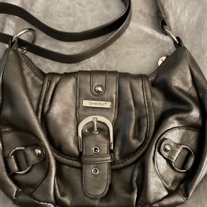 Svart axelväska från Daniel Ray - Säljer en silvrig axelväska från Daniel Ray i syntetmaterial. Väskan har dekorativa spännen, dragkedja upptill och flera fack inuti. Perfekt storlek för vardagens alla prylar och har snygga silvriga detaljer.