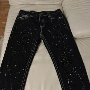 True religion jeans - Snygga svarta jeans från True Religion med vita kontrastsömmar och coola färgstänk. De har en klassisk femficksdesign och knappar med märkets logga. Perfekta för en unik stil! Aldrig använd👌