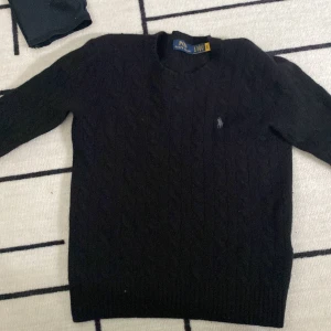 Svart kabelstickad tröja från Polo Ralph Lauren - Kabelstickad svart tröja från Polo Ralph Lauren med rund halsringning och diskret broderad logga på bröstet. Klassisk modell med ribbade muddar vid ärmslut och nederkant. Perfekt för en stilren och tidlös look.