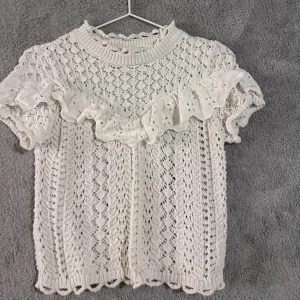 Vit stickad topp med volang - Superfin vit stickad topp med korta ärmar och dekorativ volang framtill. Sött hålmönster över hela tröjan och rund halsringning. Perfekt för dig som gillar romantisk och feminin stil.