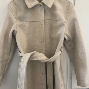 Beige kappa med bälte - Snygg beige kappa med bälte i midjan. Jackan har tryckknappar framtill och är långärmad.  Använd få gånger