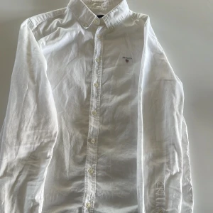 Vit skjorta från Gant - Klassisk vit skjorta från Gant med broderad logga på bröstet. Skjortan har lång ärm, knappar framtill och krage. Storlek 158/164 cm (13-14) vilket motsvarar ungefär xs i herrstorlek. Perfekt för en stilren look och passar till många tillfällen. Skriv bara vid minsta fråga, priset kan diskuteras!
