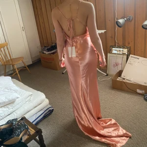 Lång rosa satinklänning med öppen rygg - Säljer en superfin långklänning i ljusrosa satin med smala axelband och öppen rygg med knyt. Klänningen har slits framtill och en mjukt draperad urringning och ett litet släp bak. Perfekt för bal eller fest! 💖Den har 2 små fläckar på släpet som knappt märks (se bilder).Orginalpriset ligger på 1399🌸