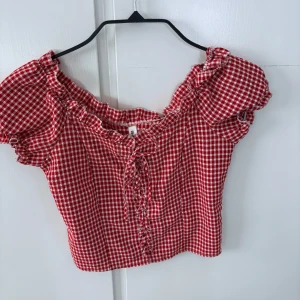 Röd och vit rutig blus med puffärmar - Supersöt röd och vit rutig blus med puffärmar och knytning framtill. Blusen har volangkant längs urringningen och är croppad i modellen. Perfekt för en somrig look!