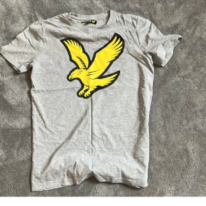 Grå t-shirt med gul fågel från Lyle & Scott - Snygg grå t-shirt från Lyle & Scott med stor gul fågel på bröstet. Perfekt för dig som gillar stilrena plagg med en cool detalj. Klassisk passform och rund hals.