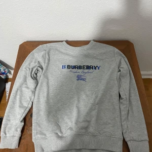 Grå sweatshirt från Burberry - Säljer en grå sweatshirt från Burberry med blå logga och text på bröstet. Tröjan har rund halsringning, långa ärmar och ribbade muddar vid ärmslut och nederkant. Perfekt för en avslappnad och stilren look.