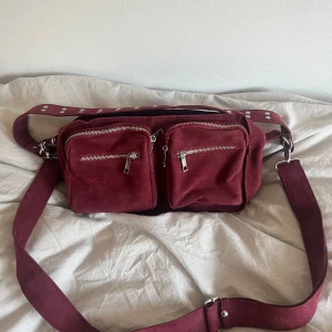 Noella väska  - Säljer en vinröd axelväska i mocka från Noella med två framfickor med dragkedja och bred axelrem med silvriga nitar. Väskan har ett stort huvudfack med dragkedja och är perfekt storlek!